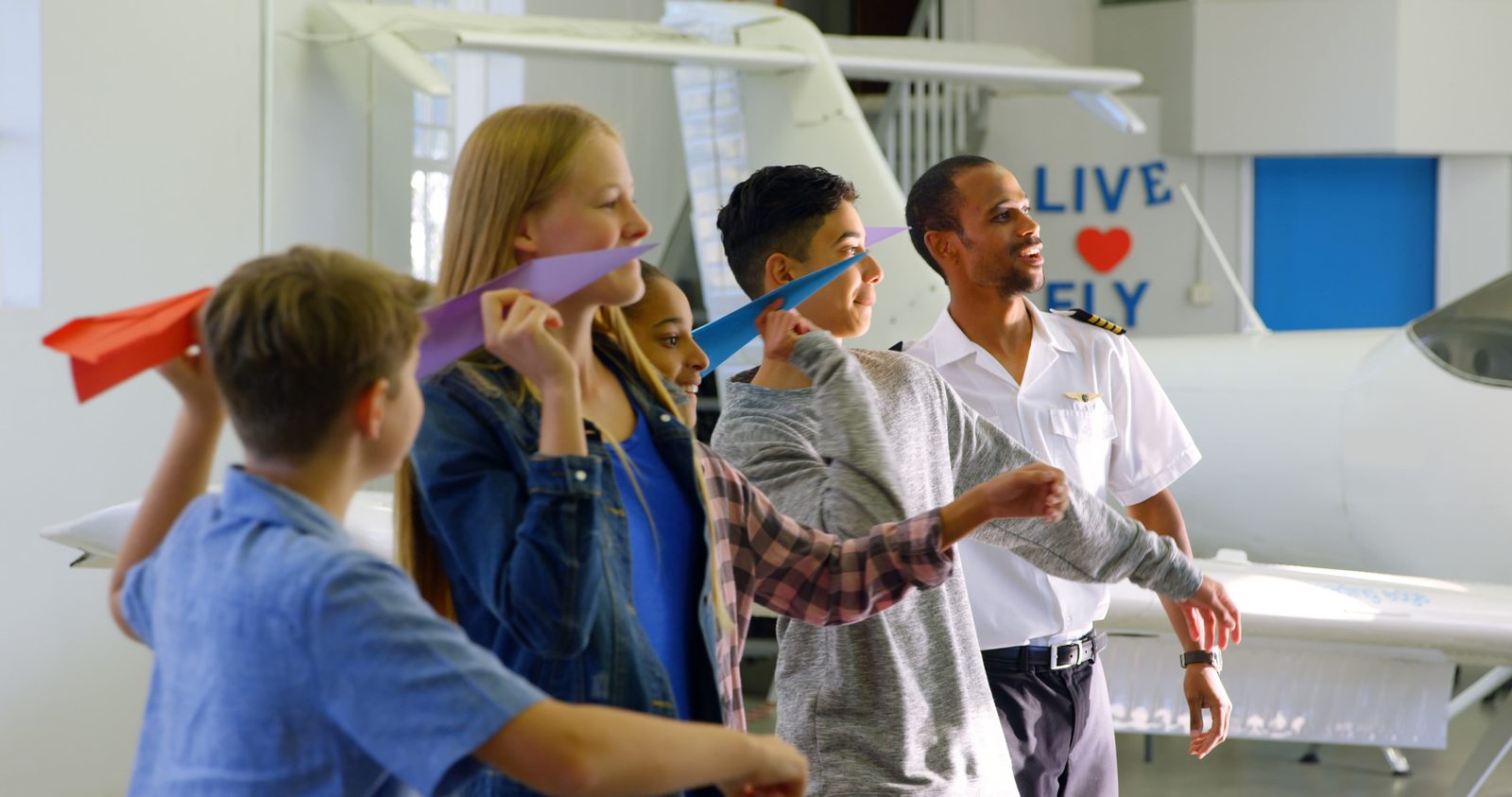 High school students throwing paper airplanes with a volunteer pilot in an aircraft hangar
